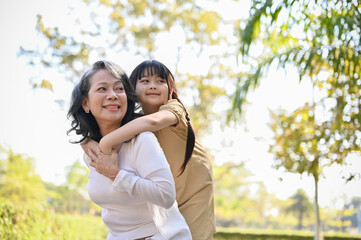 Fototapeta premium Happy Asian grandmother and her lovely granddaughter in the park, piggy back, happy family time