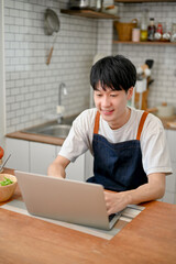 Asian man using laptop in his kitchen, joining online cooking class, searching food recipe