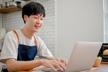 Happy Asian man in apron using laptop at his kitchen counter, searching food recipe