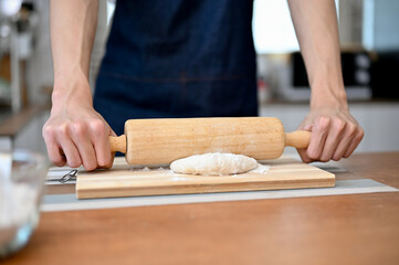 Close-up hands image of a man wearing apron holding a rolling pin, making dough on kitchen table