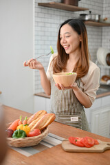 An attractive Asian woman wearing apron, holding a salad bowl, eating salad in her kitchen.