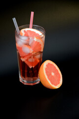 A tall glass of grapefruit juice with ice and straws on a black background, next to pieces of ripe citrus.