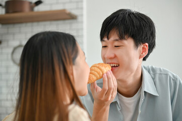 Loving Asian couple eating croissant in the kitchen together, having breakfast together