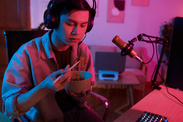Teenage boy in headphones eating ramen and watching video of blogger on computer