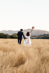 Newlywed couple walks through the countryside with bouquet in hand with the mountains in the background.