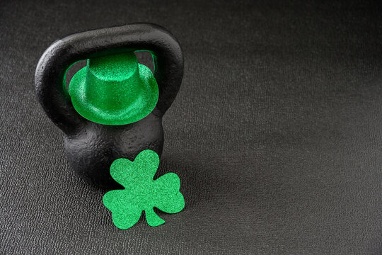 Black iron kettlebell and green glitter shamrock and leprechaun hat on a black gym floor, happy St. Patrick’s Day
