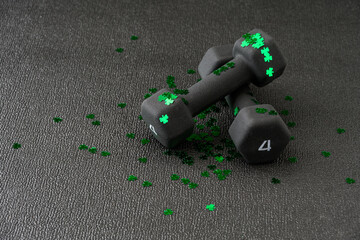 Pair of small hand weights covered in green shamrock confetti on a black gym floor, happy St....