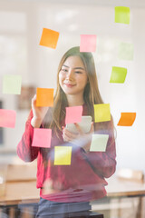 Asian businesswoman creating project plan on office wall with sticky paper notes. stylish confident manager working on business, financial and marketing planning projects. 