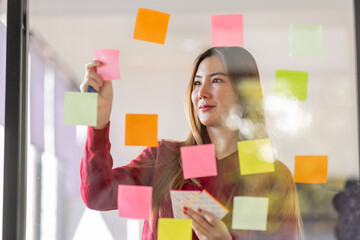 Asian businesswoman creating project plan on office wall with sticky paper notes. stylish confident manager working on business, financial and marketing planning projects. 