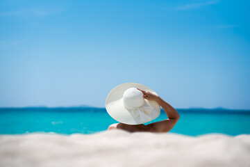 Fototapeta na wymiar A girl in a hat on the white sand on the beach. The concept of a summer vacation is a sunbathing woman in a hat enjoying the sea and the sun.