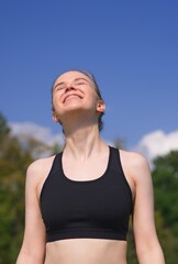 Fototapeta premium Happy positive fit fitness girl, joyful young woman is enjoying sun, good summer weather, sunbathing at sunny day on natural green and sky background. Vertical photo.