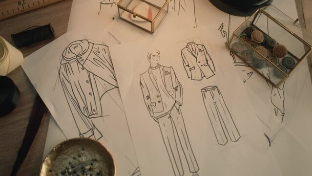 Cinematic shot of table with drawings, needles, threads, buttons, tapes, soap and ruler. Sketches of clothes, suits and jackets. Designer atelier or tailoring studio. Fashion and hand craft concept.