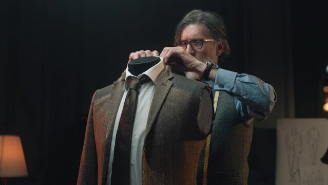 Male mature tailor works on elegant business or wedding suit. Mannequin in tailored shirt, tie and jacket in stylish luxury designer atelier or tailoring dim studio. Fashion and hand craft concept.