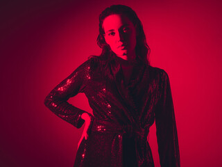 Young beautiful confident brunette female in trendy evening shiny green reflecting dress. Sexy carefree woman isolated on red neon light in studio. Fashionable model. Fantasy style. High fashion