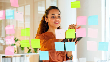 Happy female designer planning ideas on a glass wall with colorful sticky notes inside a creative...
