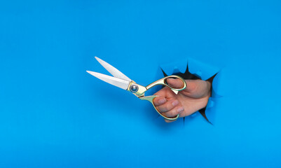 female hand torn a hole in blue paper wall and holding gold sewing scissors. Cut the price or taxes...