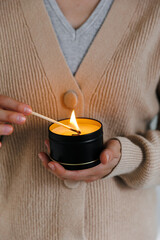 Soy scented candles in metal jars, boxes. Candle held by a girl against a sweater. A long match burns.  Modern handmade coconut wax candles without paraffin. 