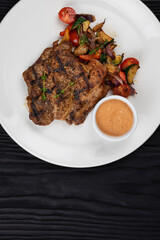 Fried pork chop with grilled vegetables and sauce on white plate on black wooden background