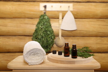 Fototapeta na wymiar Bottles of healing essential oil stand on a wooden bench next to a rosemary branches and towel against the background of sauna accessories hanging on a log wall. The concept of natural aromatherapy, s