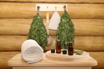 Bottles of healing essential oil stand on a wooden bench next to a towel against the background of sauna accessories hanging on a log wall. The concept of natural aromatherapy, sauna and spa treatment