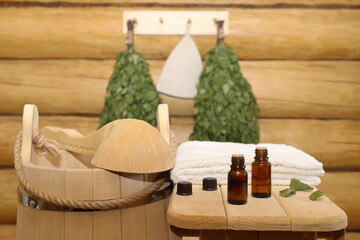 Bottles of healing essential oil stand on a wooden bench next to a towel against the background of sauna accessories hanging on a log wall. The concept of natural aromatherapy, sauna and spa treatment
