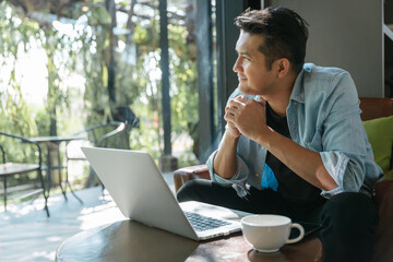 Asian handsome man sitting happily looking out of the window in a cafe. Concept freelance and financial freedom. - obrazy, fototapety, plakaty