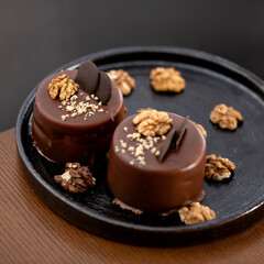 Two chocolate small desserts on plate. Cakes are decorated with nuts and chocolate glaze. Festive baking. Wooden table. View from above. Dark background. Close-up. Soft focus. Copy space. 