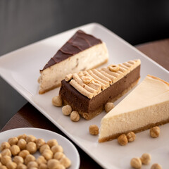 Three slices of different types of cheesecake on white rectangular plate. Dessert served with nuts on wooden table. Hazelnuts in bowl. View from above. Soft focus.