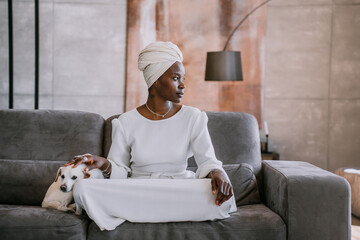 Confident African American young woman in turban sitting at home on cozy couch strokes little white...