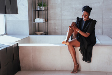 Smiley African girl in black turban and dress at bathroom putting body lotion on legs. Cheerful...