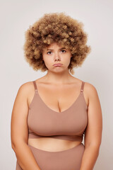 Vertical shot of upset woman in plus size dressed beige lingerie, she ot a few extra pounds for last few days, so she is looks unhappy, isolated next to white wall