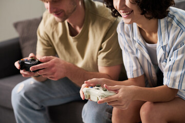 Cropped shot of young restful couple enjoying new video game while sitting next to one another and pushing buttons of joysticks