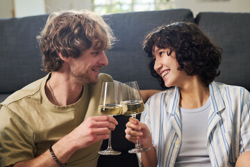 Young cheerful husband and wife clinking with flutes of champagne and looking at one another with smiles while relaxing by couch at home