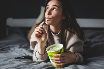 A young woman lies on her bed while eating a pint of pistachio ice cream with spoon. She is lick spoon with ice cream. Eating in bed. Happy beautiful woman resting in her comfortable bed at home.  - obrazy, fototapety, plakaty