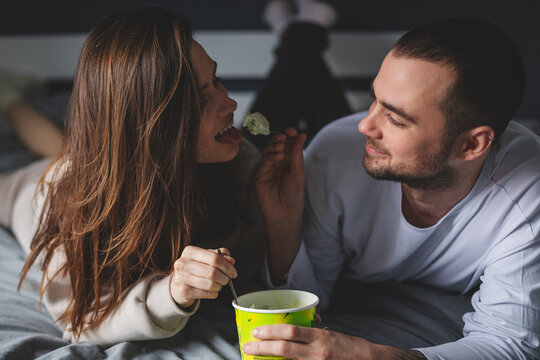 Cheerful Couple Eating Pistachio Ice Cream Lying In The Bed At Home. Guy Feeding His Girlfriend With Spoon. Dating. Couple Have Fun At Home.