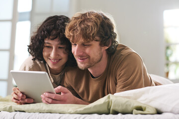 Happy young couple with tablet watching amusing online video or movie while staying in bed after sleep in the morning and enjoying rest