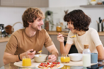 Happy young man and his wife eating cornflakes with milk for breakfast while sitting by kitchen table served with sandwiches and juice