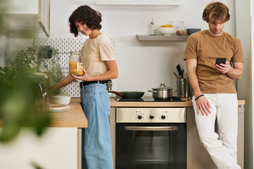 Young woman holding jar of cornflakes while standing by sink and her husband with smartphone...