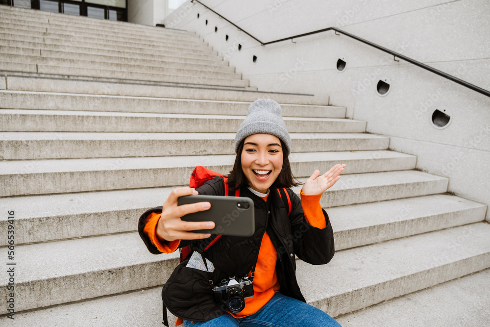 Wall mural Smiling asian woman taking selfie and gesturing while sitting on stairs