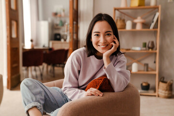 Beautiful asian woman smiling at camera while sitting on armchair at home