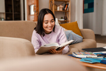 Cheerful asian woman reading book while lying on couch at home