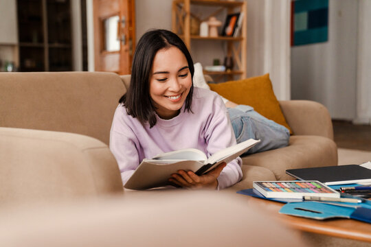 Cheerful Asian Woman Reading Book While Lying On Couch At Home