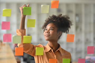 Smiling African American female manager putting sticker notes to share idea for startup project on glass board in office