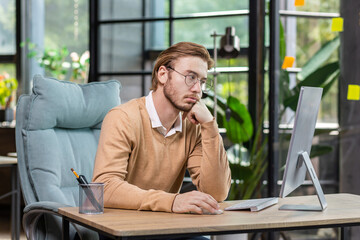 Frustrated and upset businessman reading news online from computer monitor, young blond man sad at...