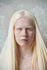 Fototapeta premium Young serene albino woman with white cover on head standing in front of camera against marble wall during photo session in studio
