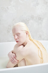 Peaceful albino girl with wet long hair sitting in warm water in white bathtub and embracing herself while enjoying relaxation procedure - obrazy, fototapety, plakaty
