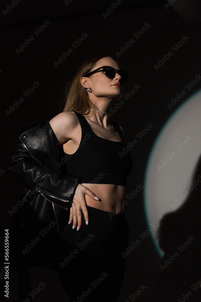 Sticker Cool fashion hipster beautiful woman with sunglasses in Stylish black top and fashionable leather jacket poses in the dark in studio