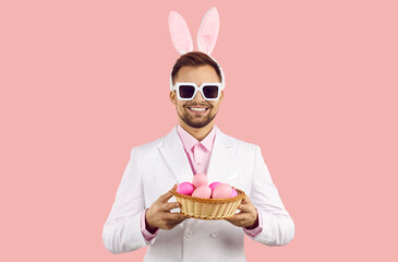 Portrait of smiling man in bunny ears hold box of painted eggs celebrate Easter. Happy male in rabbit costume isolated on pink studio background with basket on egg hunt. Celebration concept.