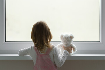 Little girl holding white teddy bear and standing alone at window and looking out from home. Back...