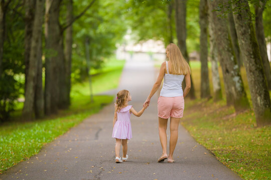 Little Daughter And Young Adult Mother Speaking And Walking On Sidewalk Through Tree Alley At City Park. Spending Time Together In Beautiful Warm Sunny Summer Day. Back View.